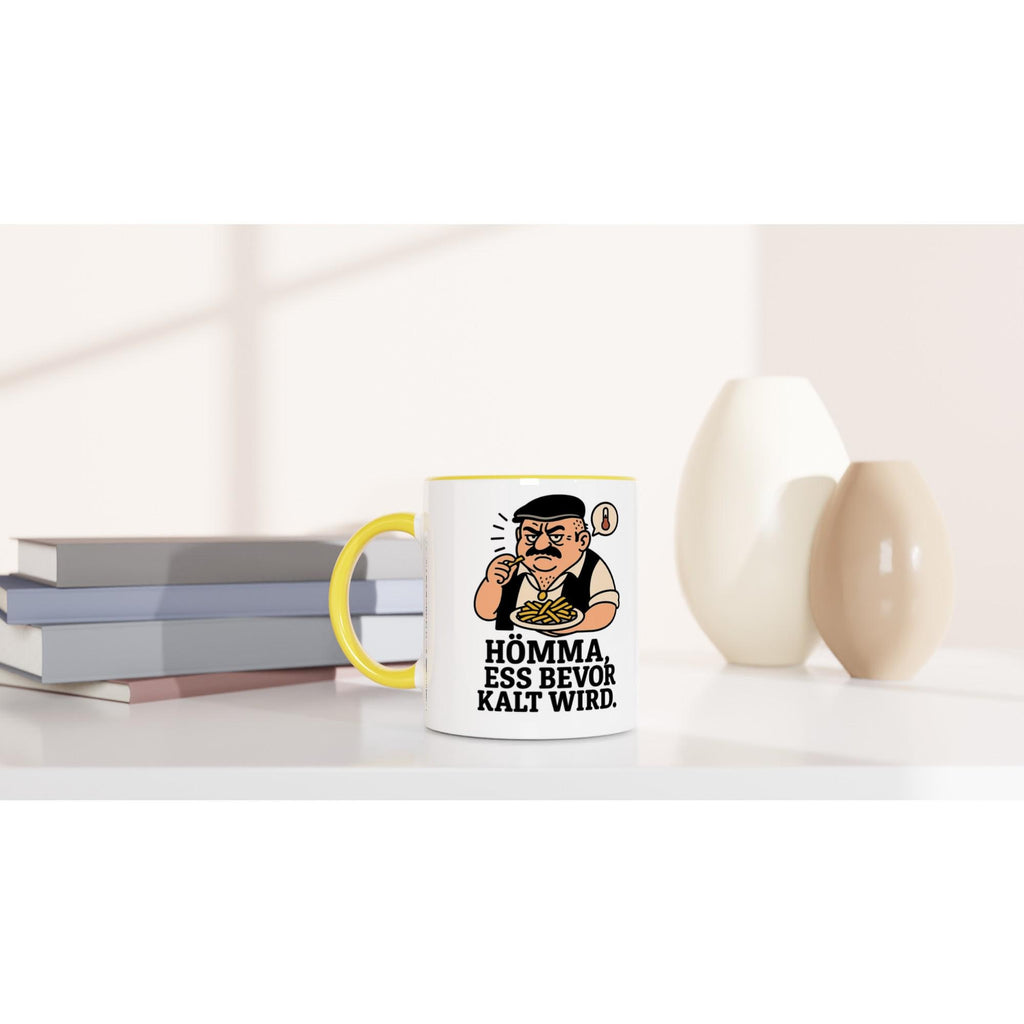 Keramiktasse mit Spruch Geschenk Homma ess bevor kalt wird mit gelbem Henkel auf weißem Tisch